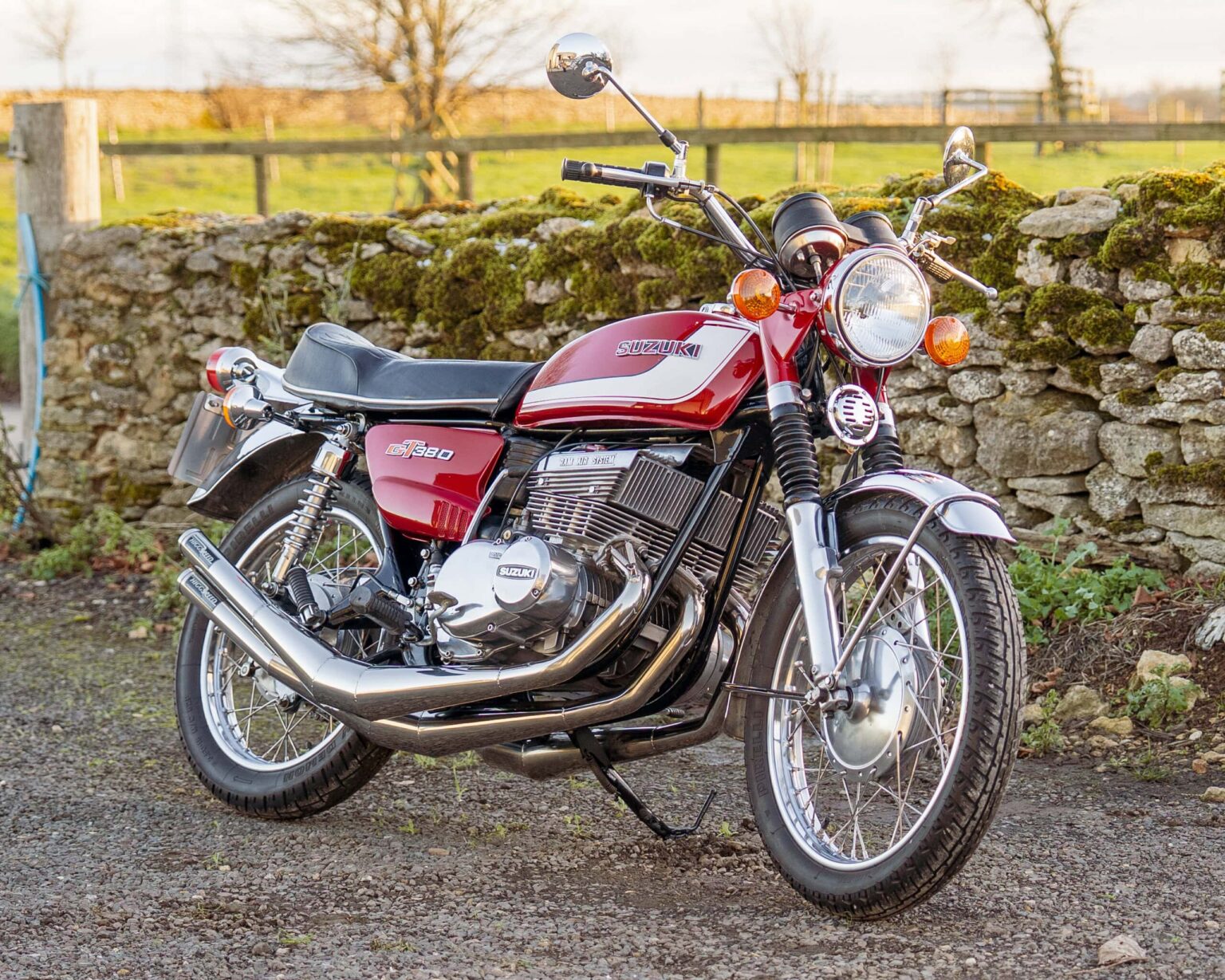 A vintage red Suzuki Gt750 motorcycle parked outdoors by a stone wall.