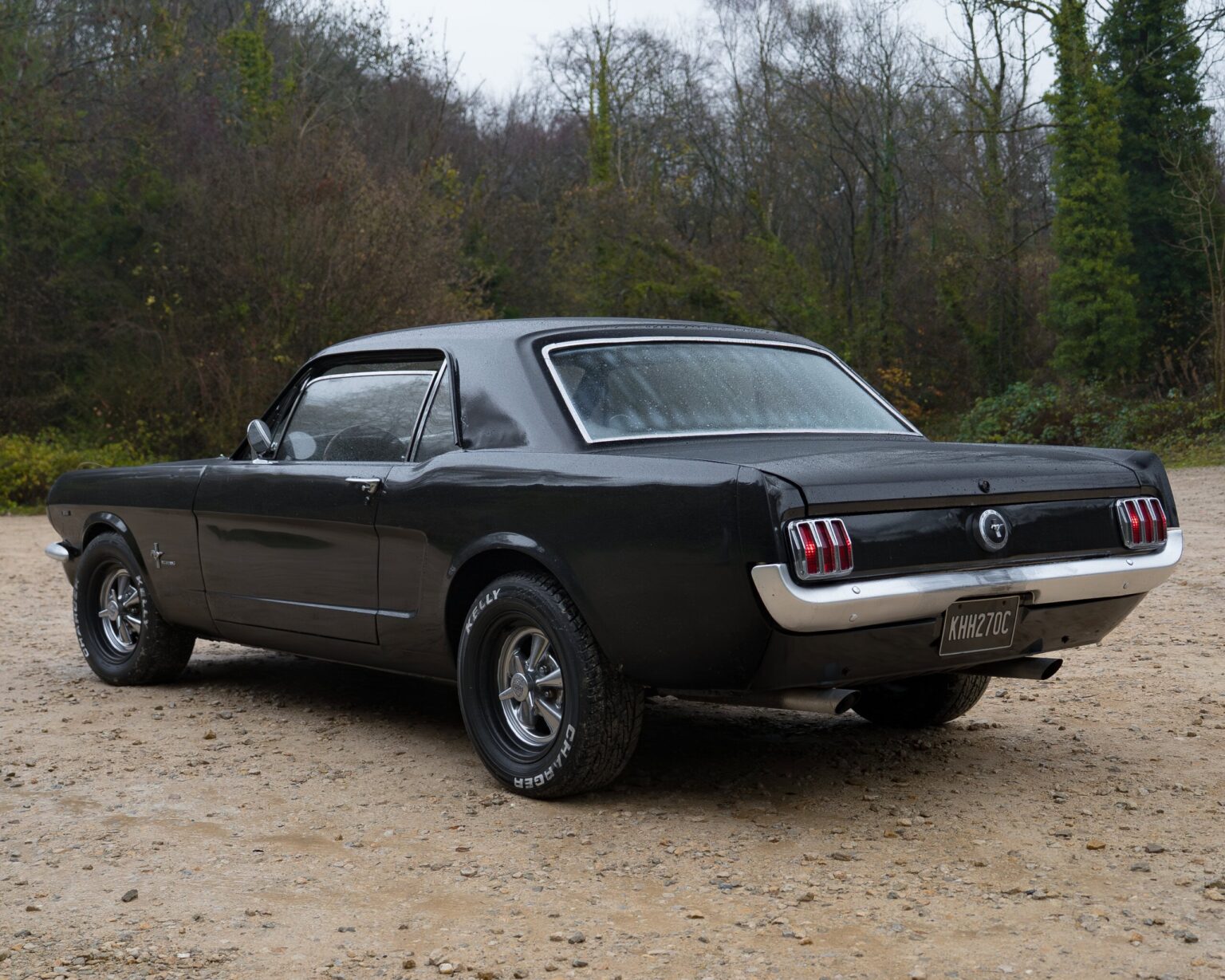 Vintage black Ford Mustang with classic design, parked outdoors on a gravel surface amid trees.