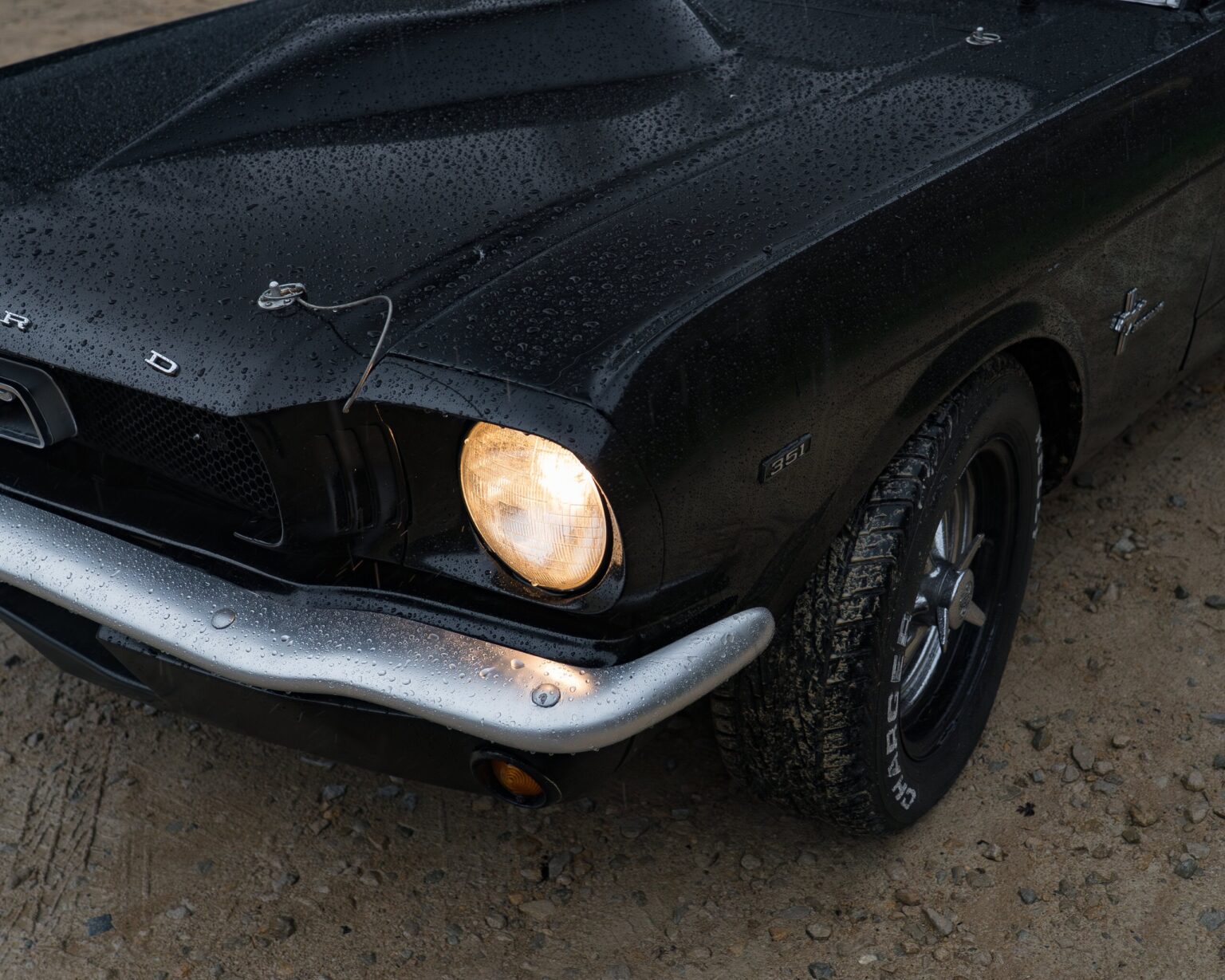 Vintage black Ford Mustang with "351" badge, rain-drenched, parked on gravel.