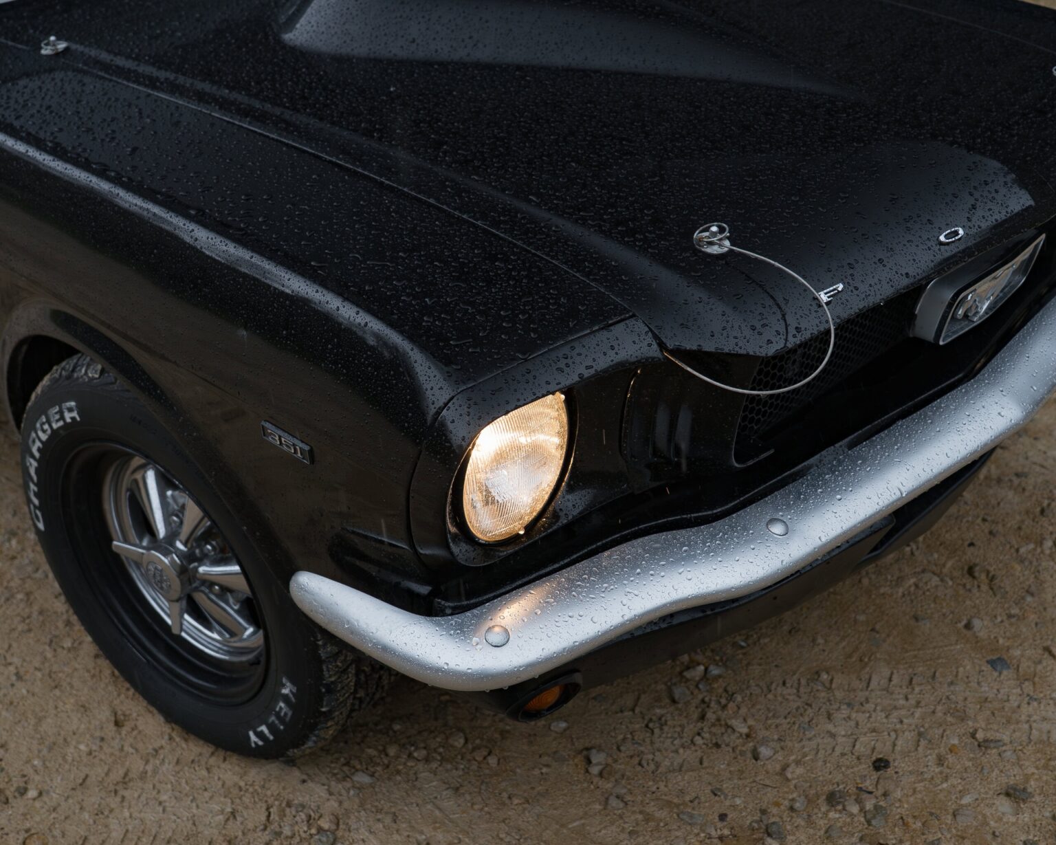 Close-up of a wet black vintage muscle car’s front with illuminated headlight and water droplets.