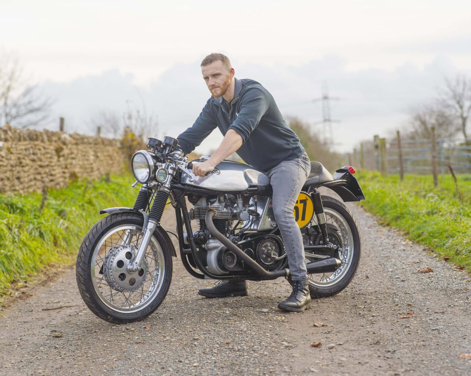 Rider sitting on a vintage motorcycle on a rural gravel path with countryside scenery.