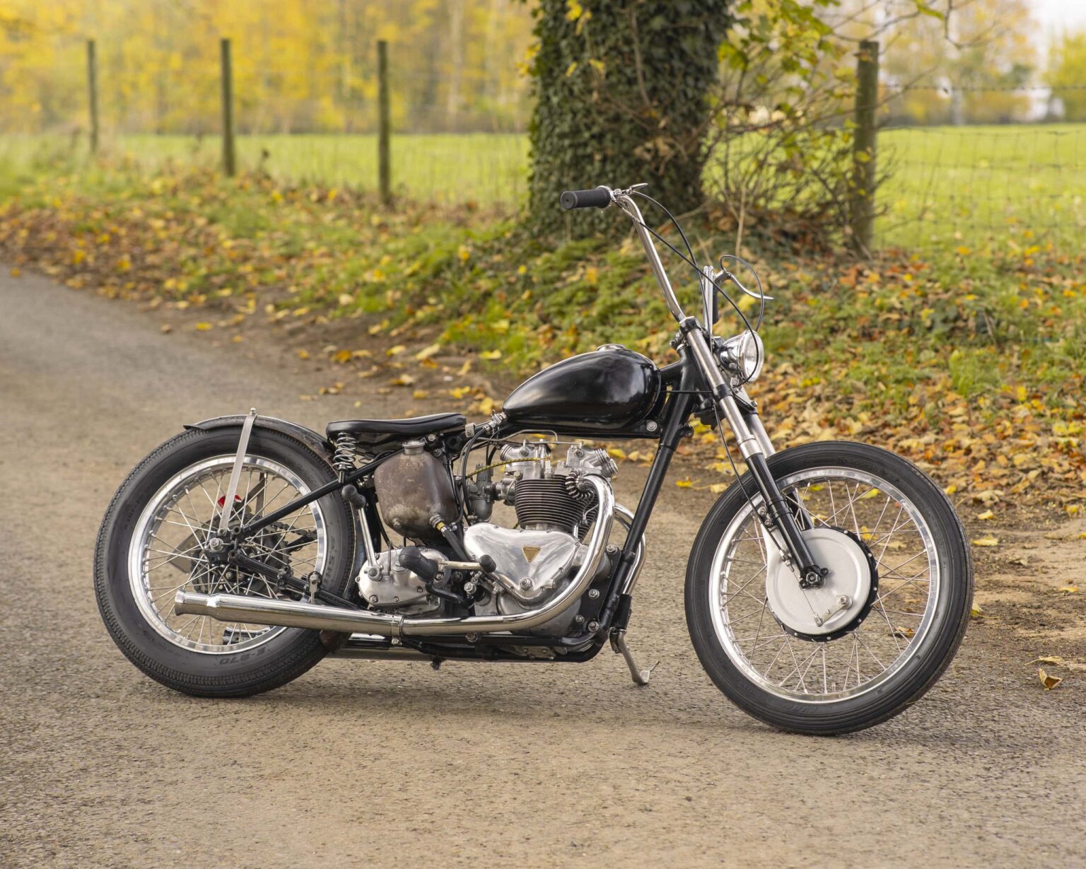 Vintage black motorcycle parked on a gravel path with fallen leaves and fall foliage backdrop.