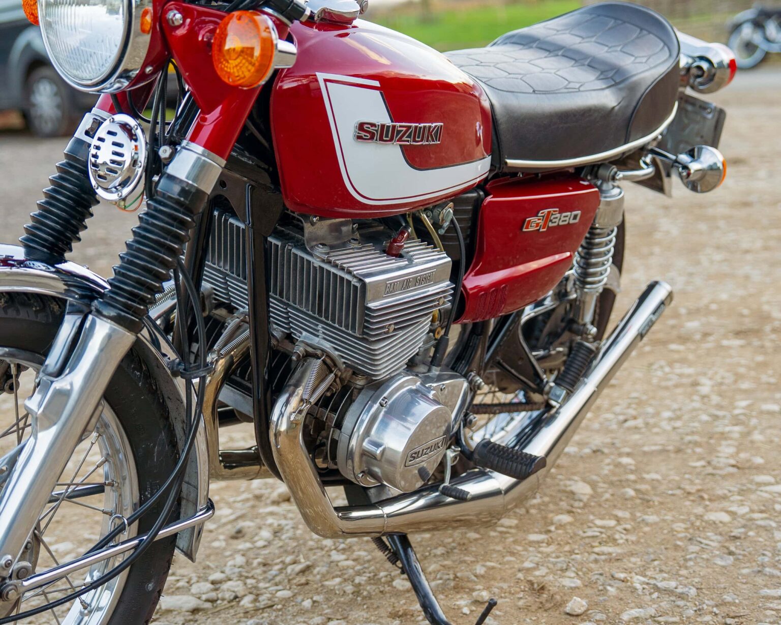 Vintage red Suzuki GT380 motorcycle with chrome details and black seat, parked outdoors at a classic car event.