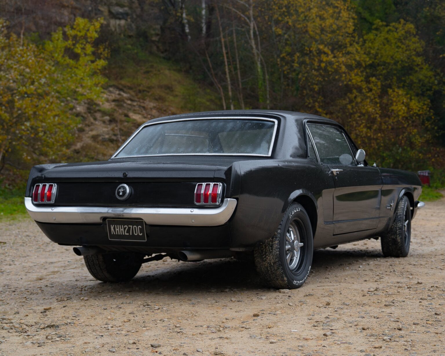 A vintage black Ford Mustang parked on a gravel road with autumn trees in the background.