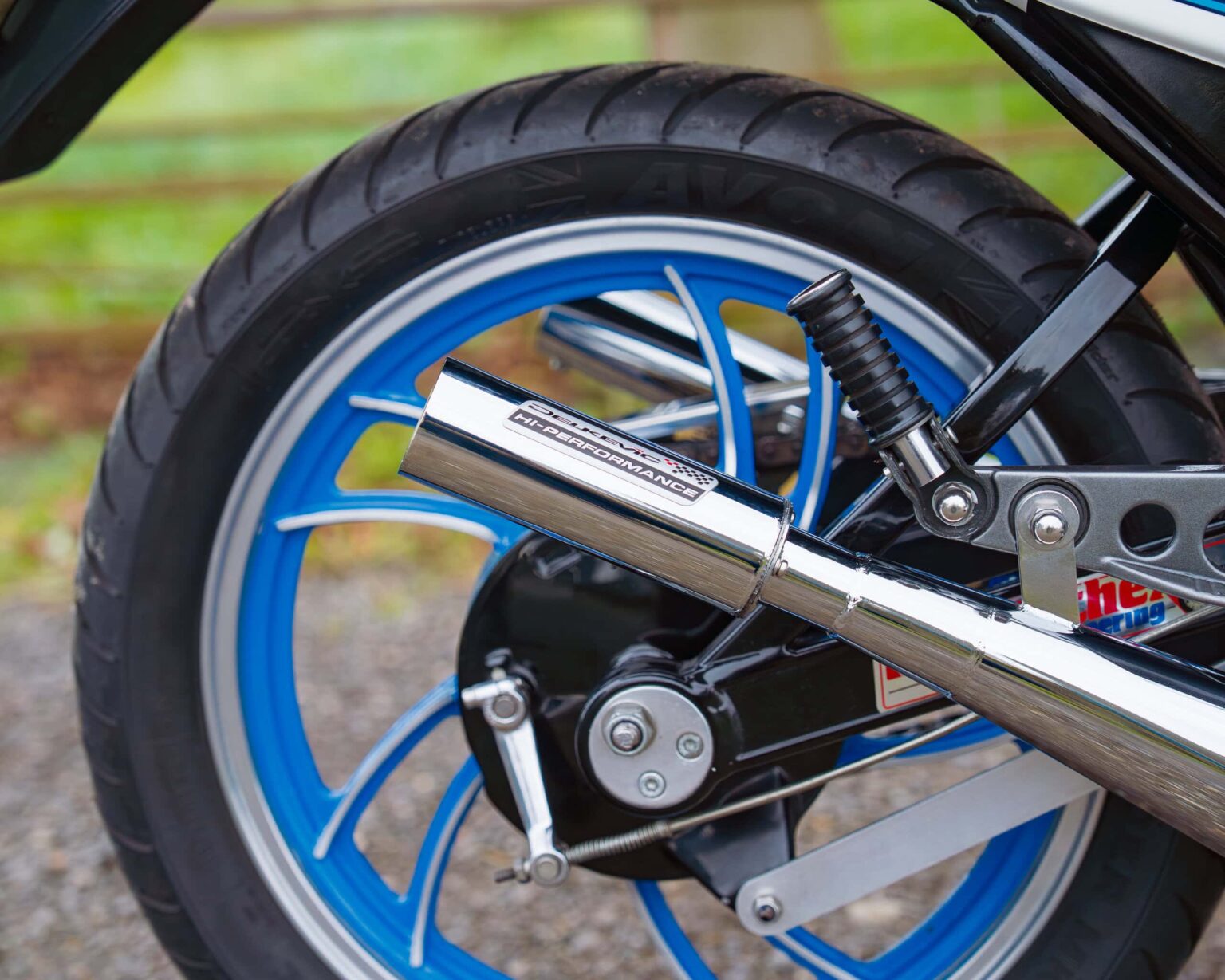 Close-up of a blue-rimmed motorcycle wheel, highlighting the high-performance exhaust and rear suspension details.