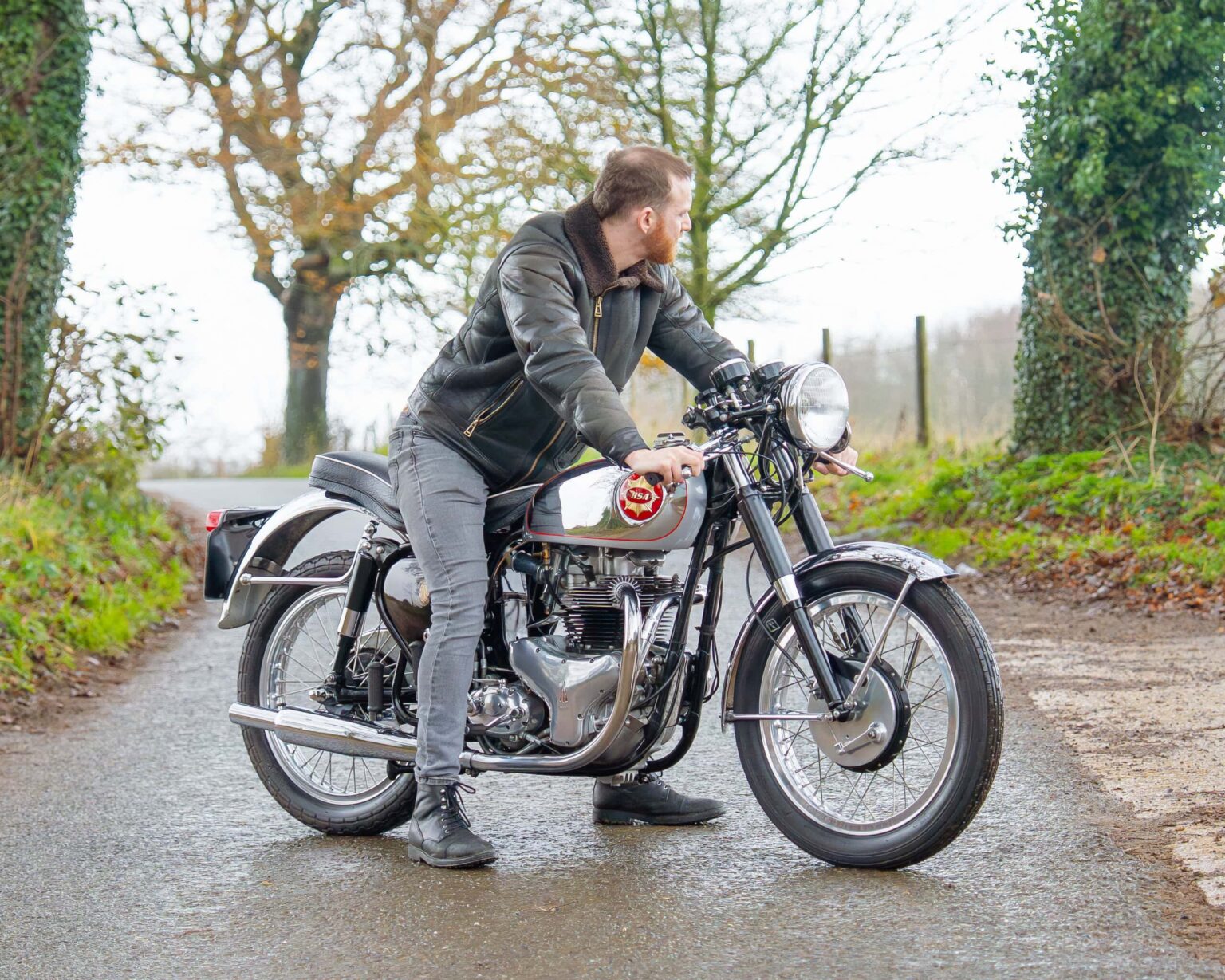 Alt text: Man in leather jacket riding a classic vintage motorcycle on a countryside road.
