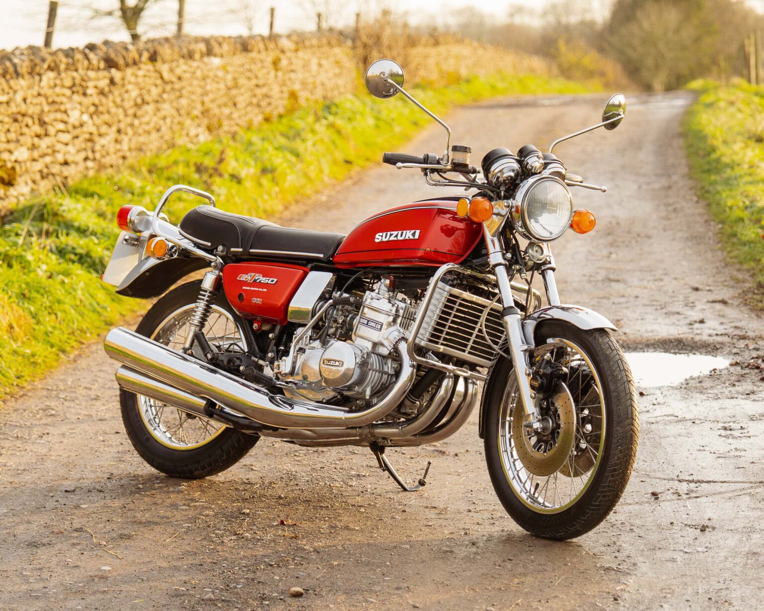 A vintage red Suzuki GT750 motorcycle parked on a rural dirt track with lush greenery and stone wall in background.