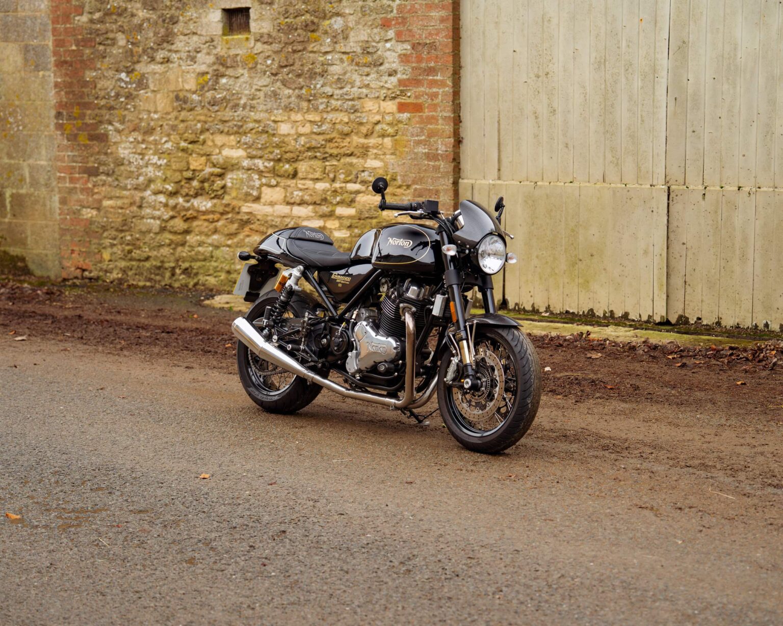 Motorcycle parked on a rural road against a brick and wooden wall.