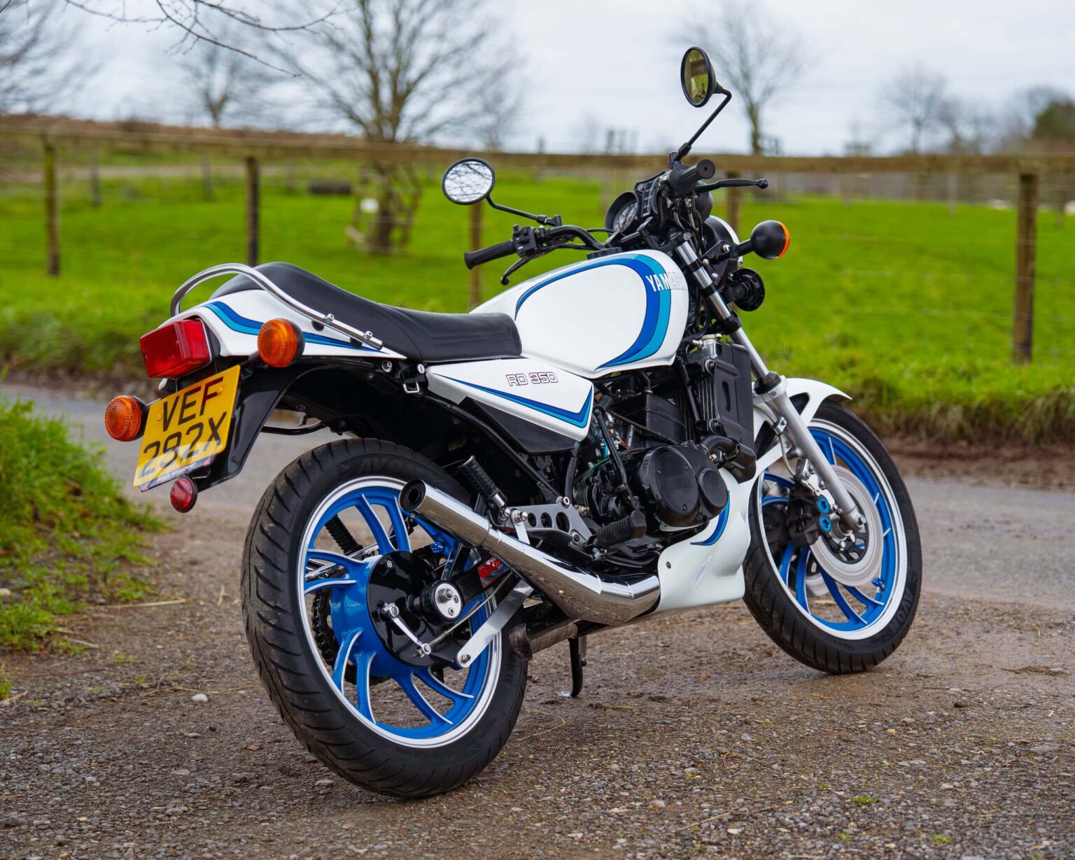VINTAGE Yamaha RD350 motorcycle on gravel with green field background.