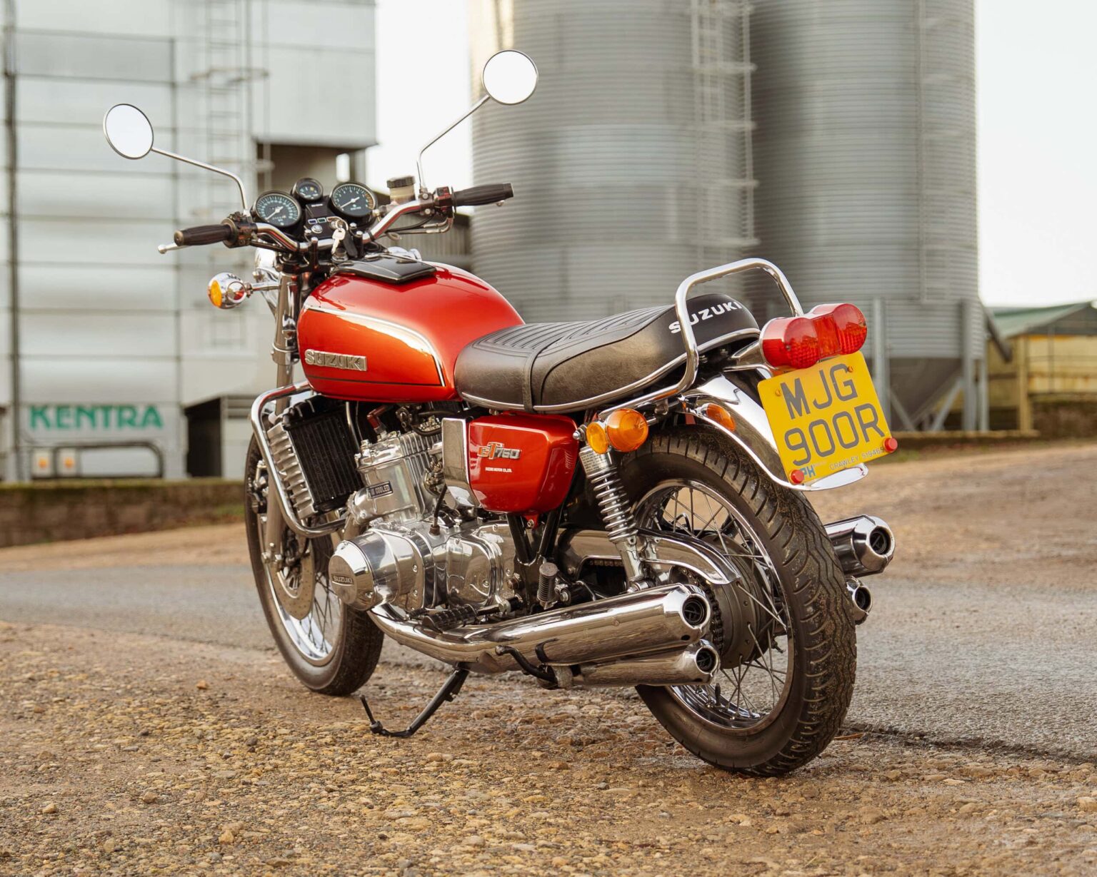 Iconic red Suzuki motorcycle with chrome highlights, vintage style, parked on gravel near industrial facilities.