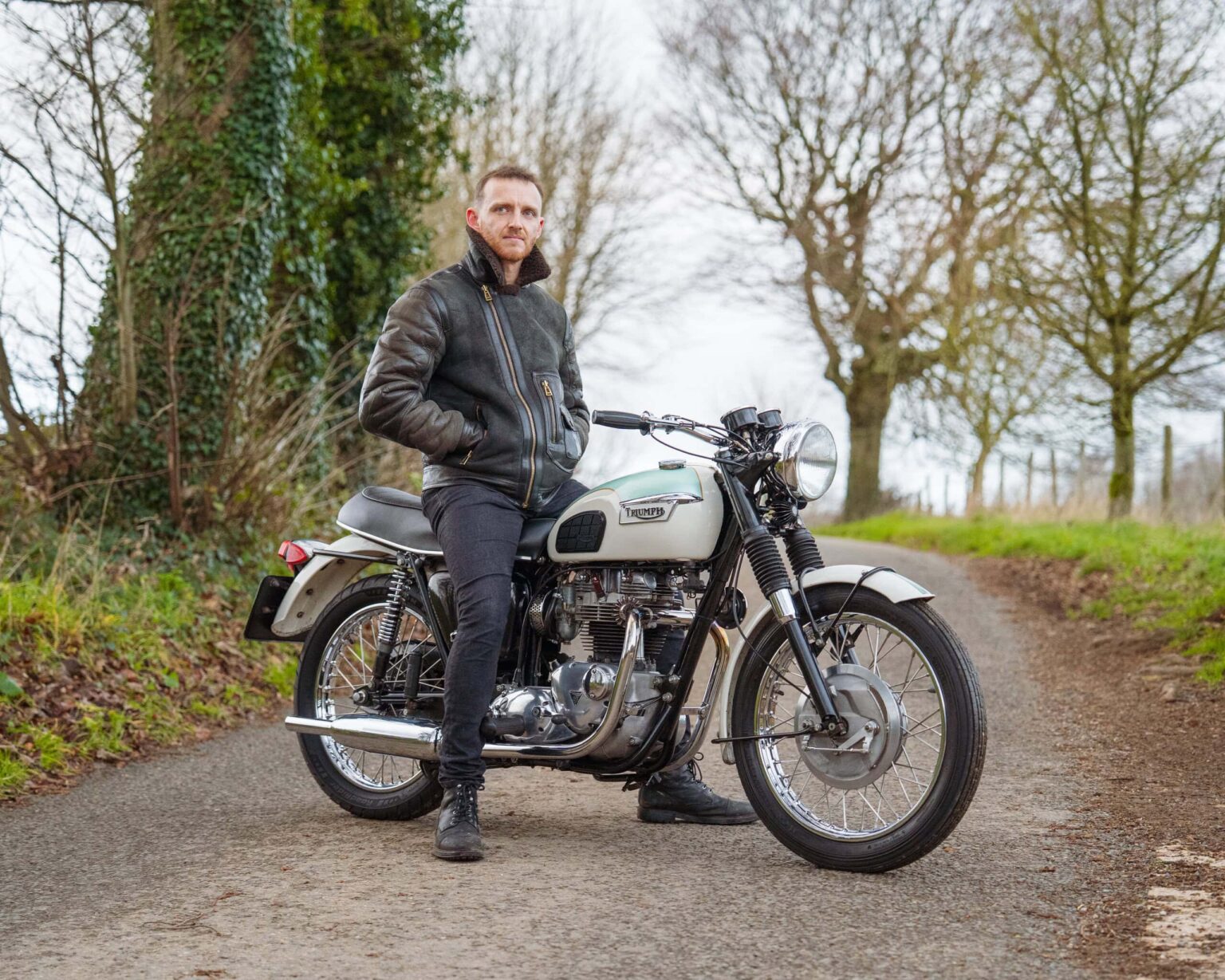 Motorcycle rider standing next to a vintage motorcycle on a rural path.