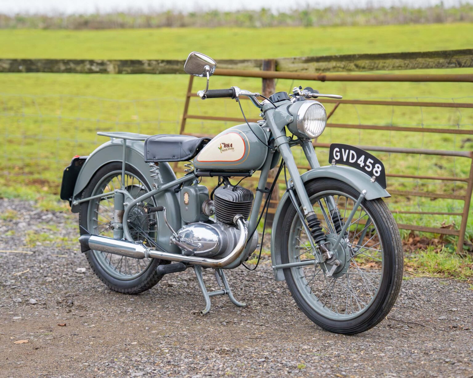 Classic motorcycle with a grey and cream colour scheme, parked outdoors near a wooden fence.